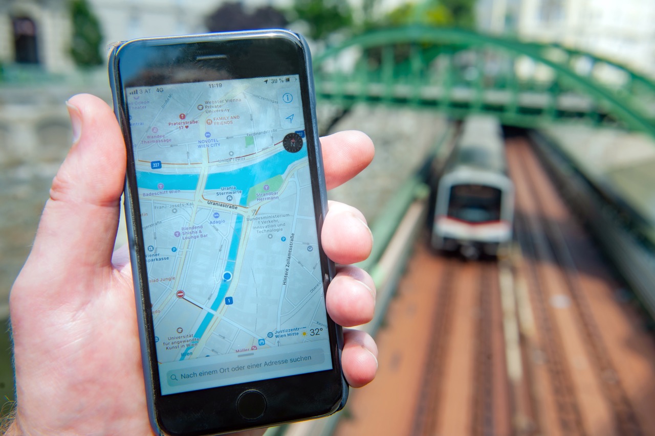 Digitale Konzeption für den Wien-Plan Eine linke Männerhand hält ein Handy, auf dem ein Ausschnitt eines Wien-Plans zu sehen ist; dahinter verschwommen Gleise mit U-Bahn und einer Brücker.