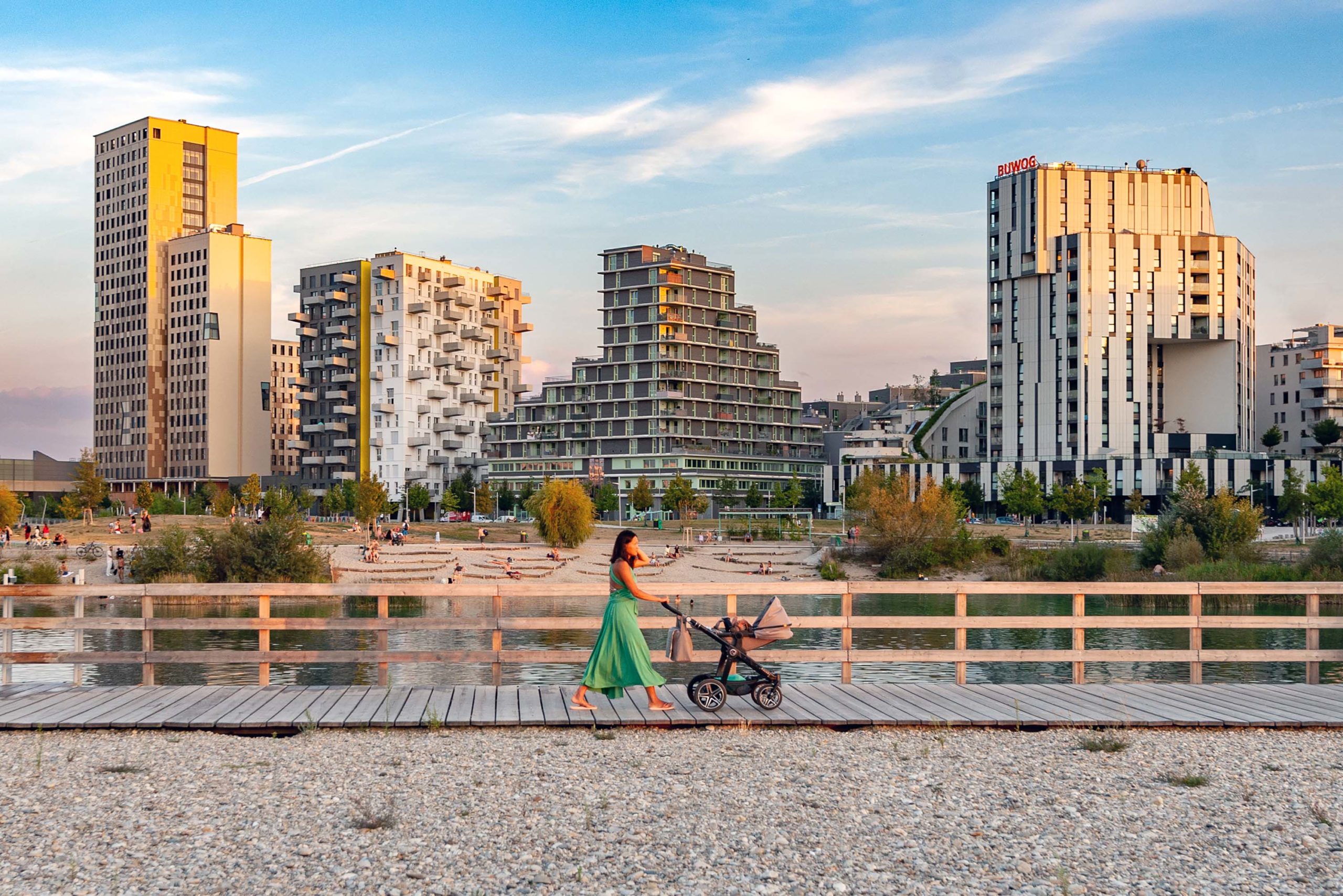 seestadt_88 Großaufnahme der Seestadt Aspern. Im Vordergrund geht eine Frau im grünen Kleid mit einem Kinderwagen am Steg neben dem Teich spazieren. © Stadt Wien / Gerd Götzenbrucker
