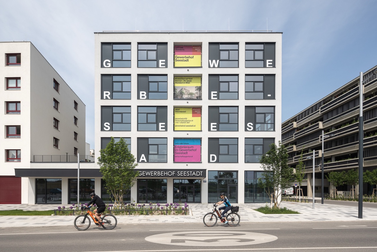 Aufnahme der Front des Gewerbehofs Seestadt, davor Grünflächen und Radfahrer*innen in einer Tempe-30-Zone.