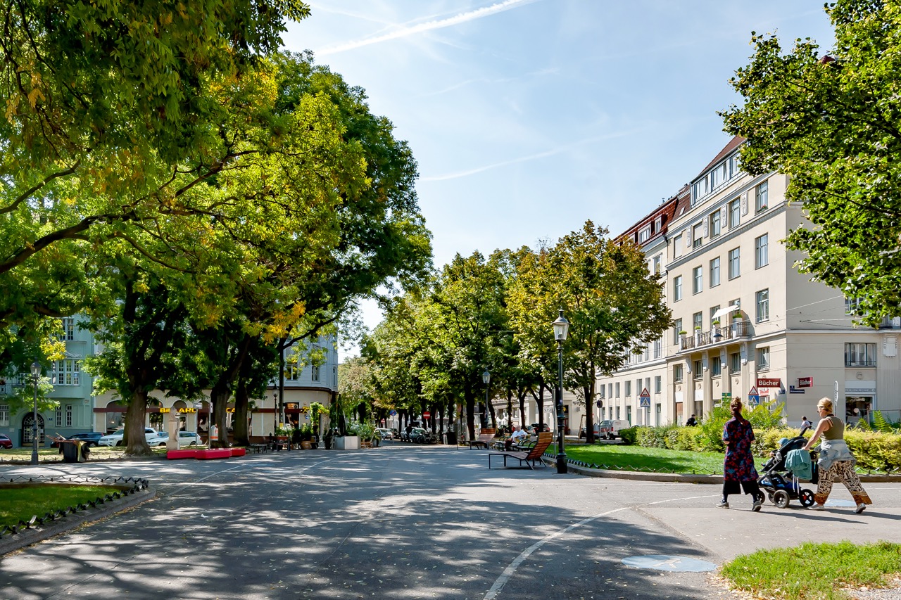 Aufnahme der begrünten Markgraf-Rüdiger-Straße im 15. Bezirk mit schattigen Bäumen, Grünflächen und Sitzgelegenheiten. Menschen, die darüber spazieren oder auf einer Bank sitzen sind klein im Bild.