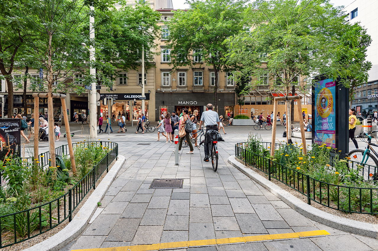 Aufnahme einer belebten, begrünten Straße in Wien mit Platz für alle Verkehrsteilnehmer*innen.