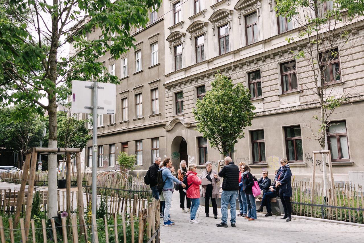 Eine Gruppe von Teilnehmer*innen bei einer Gemma-Zukunft-Tour 2025 mit Franziska Leeb steht vor Gründerzeithäusern. Der Straßenraum ist begrünt mit Bäumen und Grünbeeten, der Boden hell gepflaster.