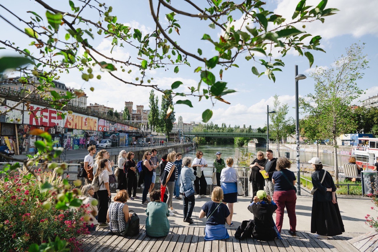 Eine Gruppe Teilnehmer*innen beim Frauen-bauen-Stadt-Spaziergang aus der Gemma-Zukunft-Reihe am Donaukanal. Im Vordergrund Sitzgelegenheit, im Hintergrund der Kanal + Ufer